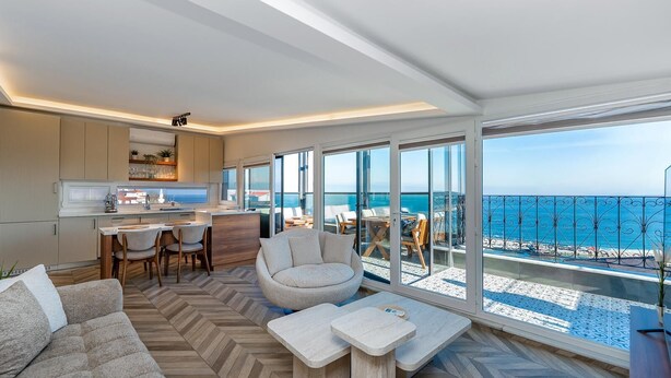 A modern living room with a sofa, coffee table, and dining area, overlooking a balcony with a sea view.