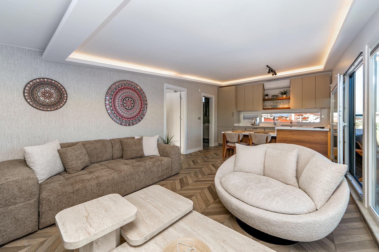 Un salon moderne avec un canapé, un fauteuil et une table basse, une cuisine avec un coin repas, et de grandes fenêtres.