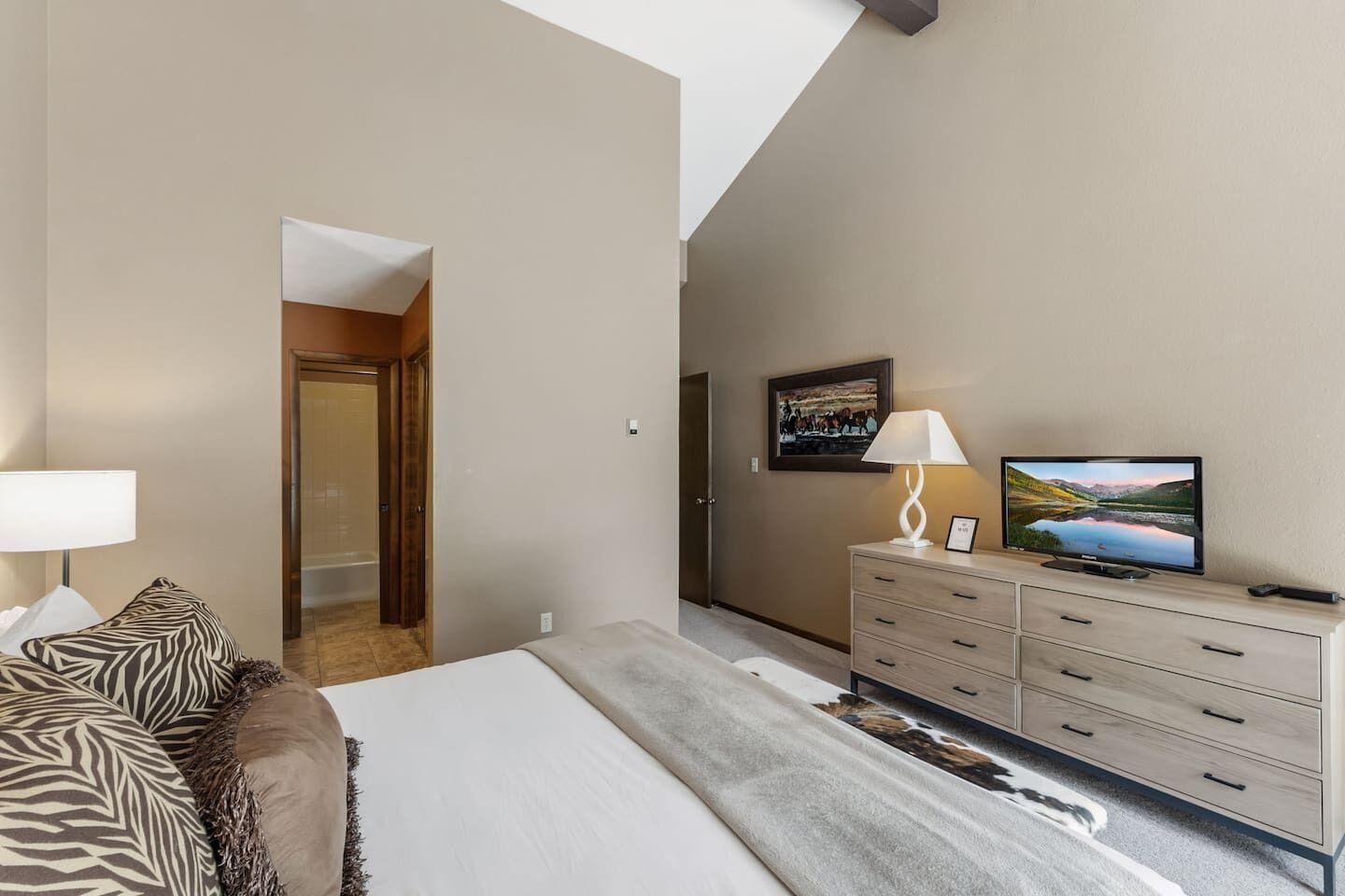 A hotel room with a bed, a TV, a dresser, and a painting on the wall.