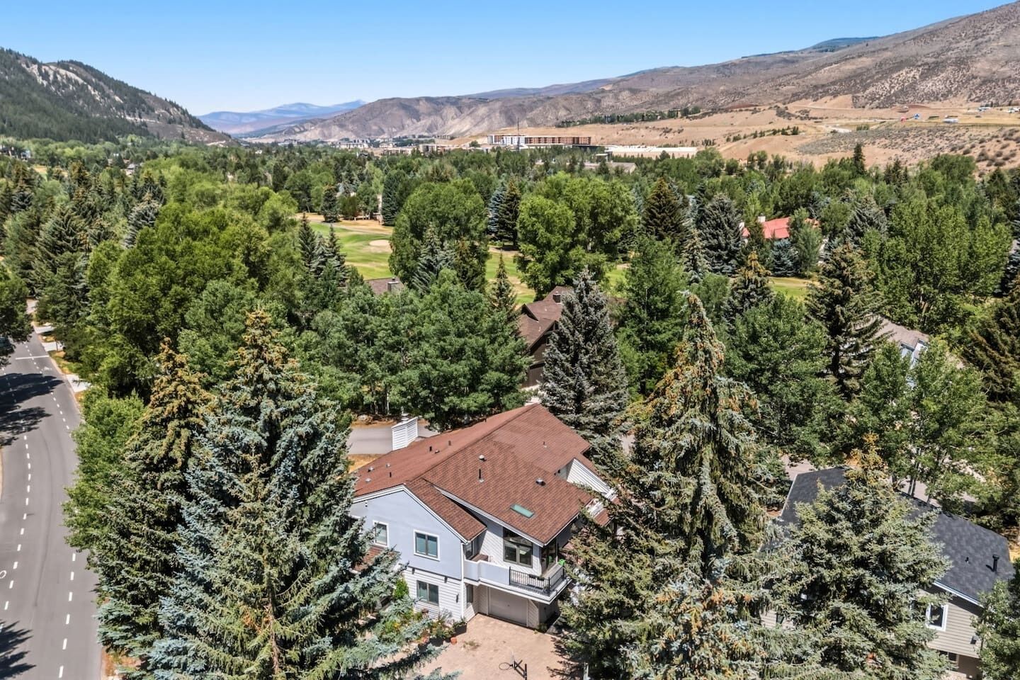 A residential area with houses surrounded by dense coniferous trees, a clear road, and a hilly landscape in the background.