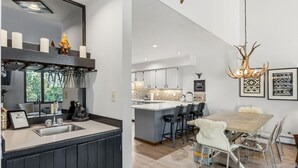 Uma cozinha moderna com uma ilha grande, uma sala de jantar com mesa e cadeiras de madeira, e um lustre com chifres de veado.