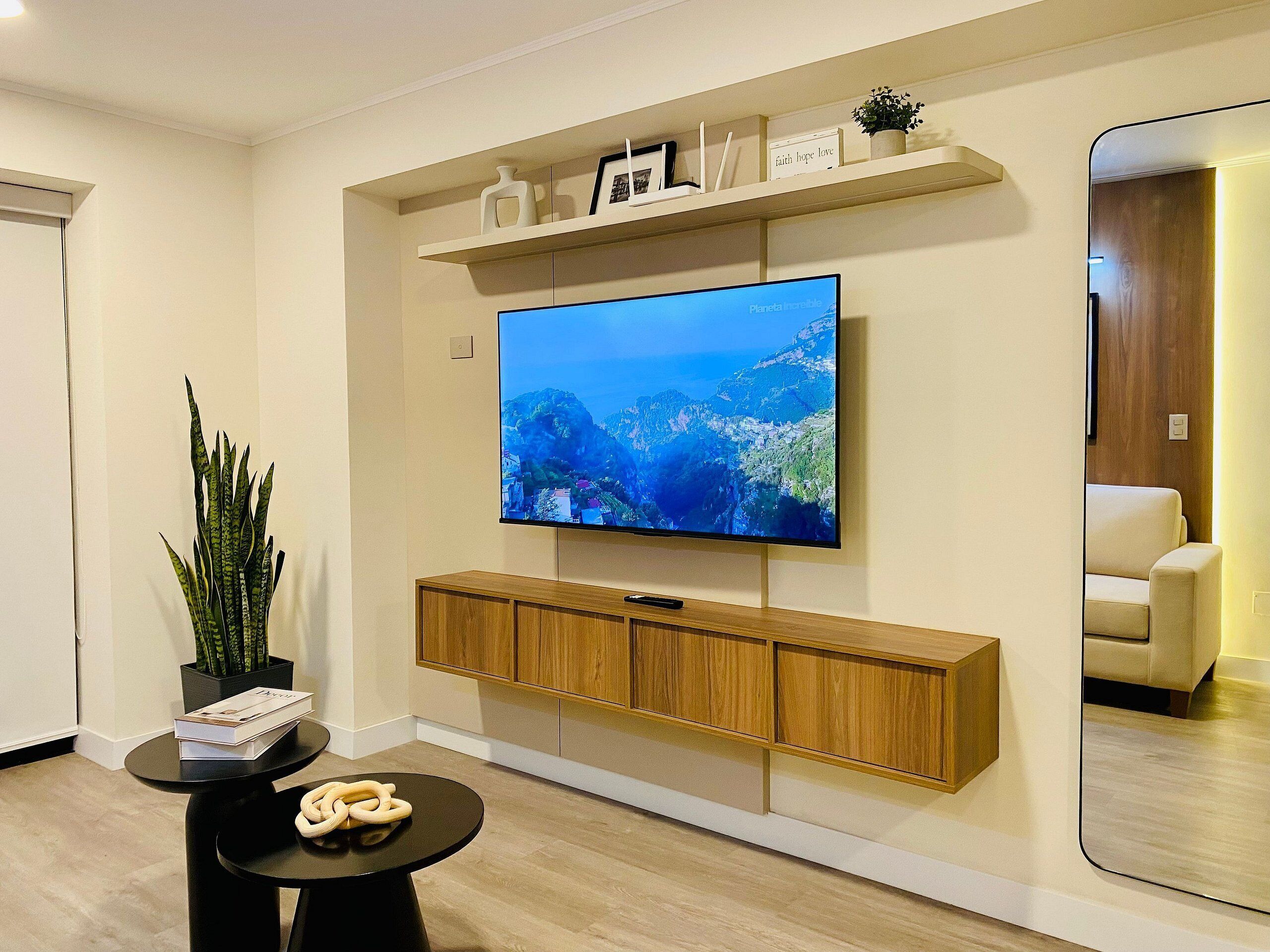 Un salon moderne avec un téléviseur à écran plat fixé au mur, des meubles en bois en dessous, une petite table ronde agrémentée d’une plante et de livres, et un grand miroir qui reflète la pièce.