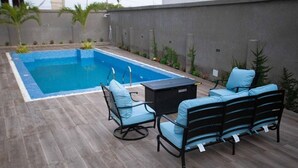 Una piscina con muelle de madera, asientos exteriores acolchados en azul y una mesa pequeña.