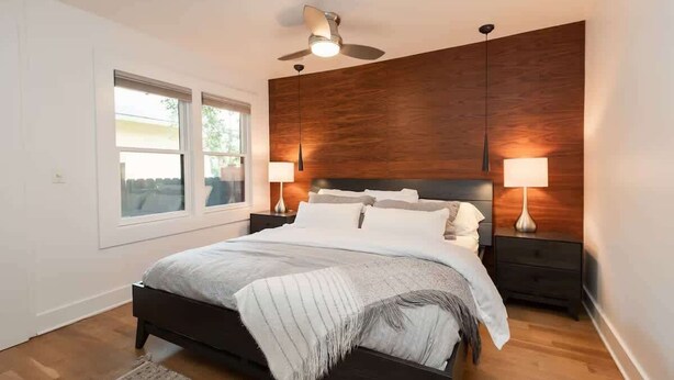 A bedroom with a bed, a ceiling fan, two bedside lamps, a window, and a wooden headboard. - The Modern Cottage Bend (Bend)