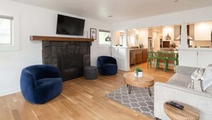 A modern living room with a fireplace, two blue armchairs, a grey sofa, a round wooden coffee table, and a TV mounted above the fireplace. - The Modern Cottage Bend (Bend)