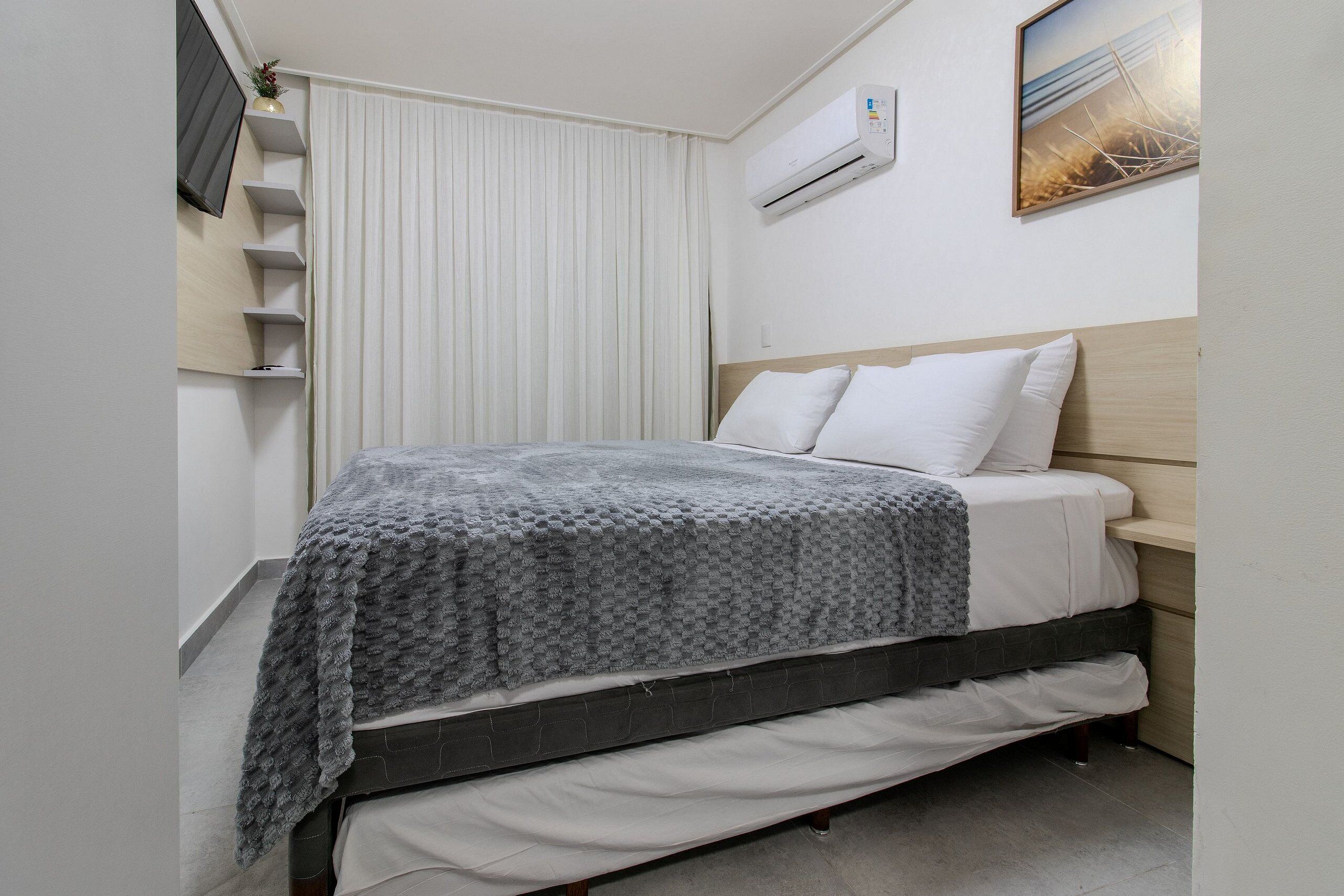 A bedroom with a bed, a bedside table, a wall-mounted air conditioner, and a window with blinds.