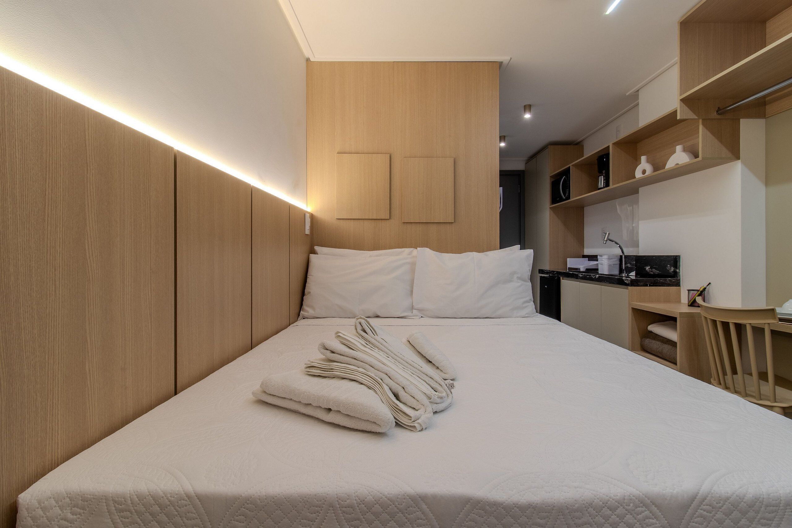 A modern hotel room with a neatly made bed, wooden paneling, a small kitchenette, and built-in shelving.