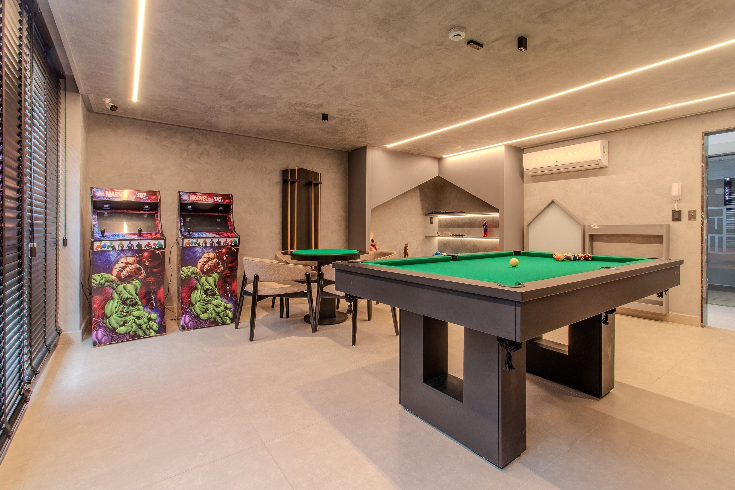 Ein Billardtisch mit grüner Spielfläche, zwei Spielautomaten mit farbenfroher Grafik, ein Sofa und ein modernes Decken-Design.