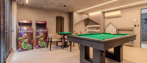 Ein Billardtisch mit grüner Spielfläche, zwei Spielautomaten mit farbenfroher Grafik, ein Sofa und ein modernes Decken-Design.