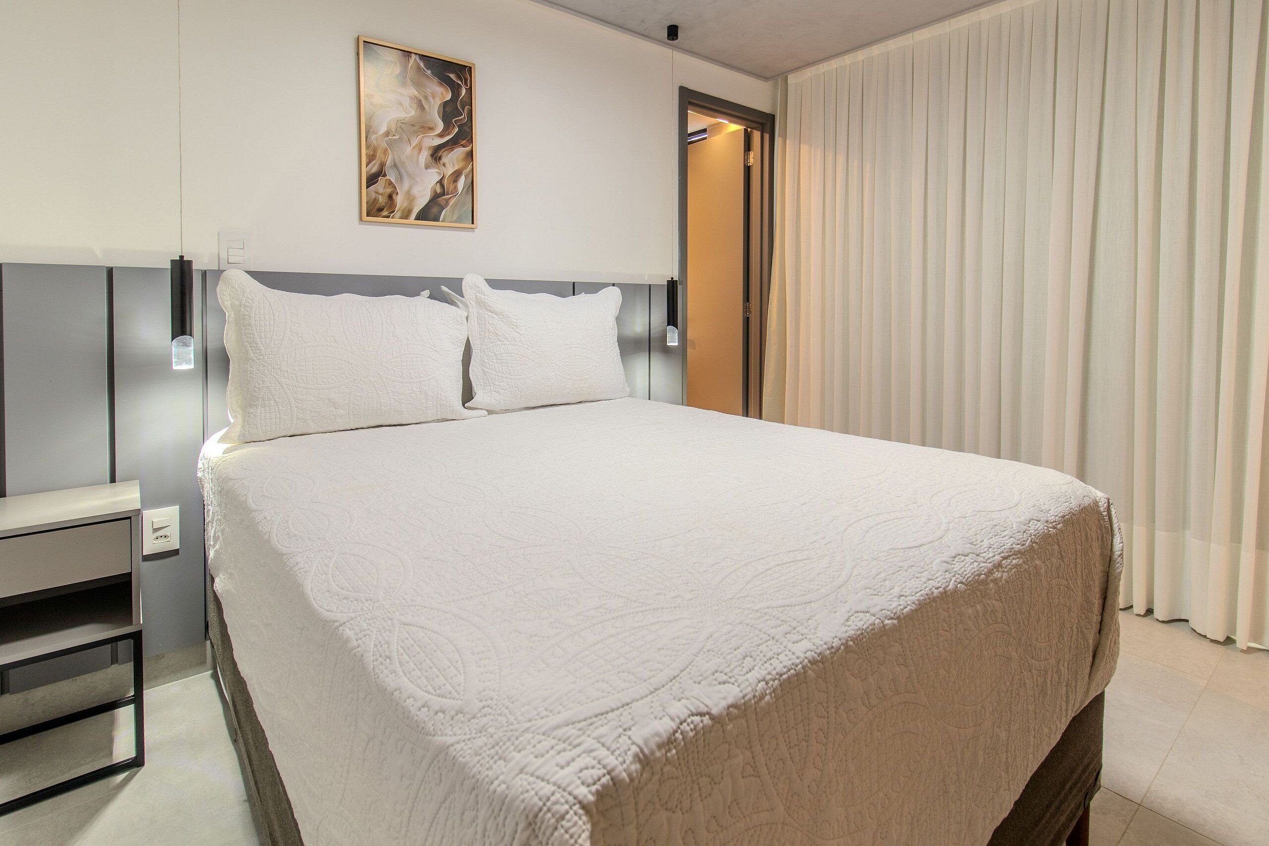 Una habitación de hotel con una cama grande, una mesita de noche, un cuadro en la pared y una puerta con cortinas.