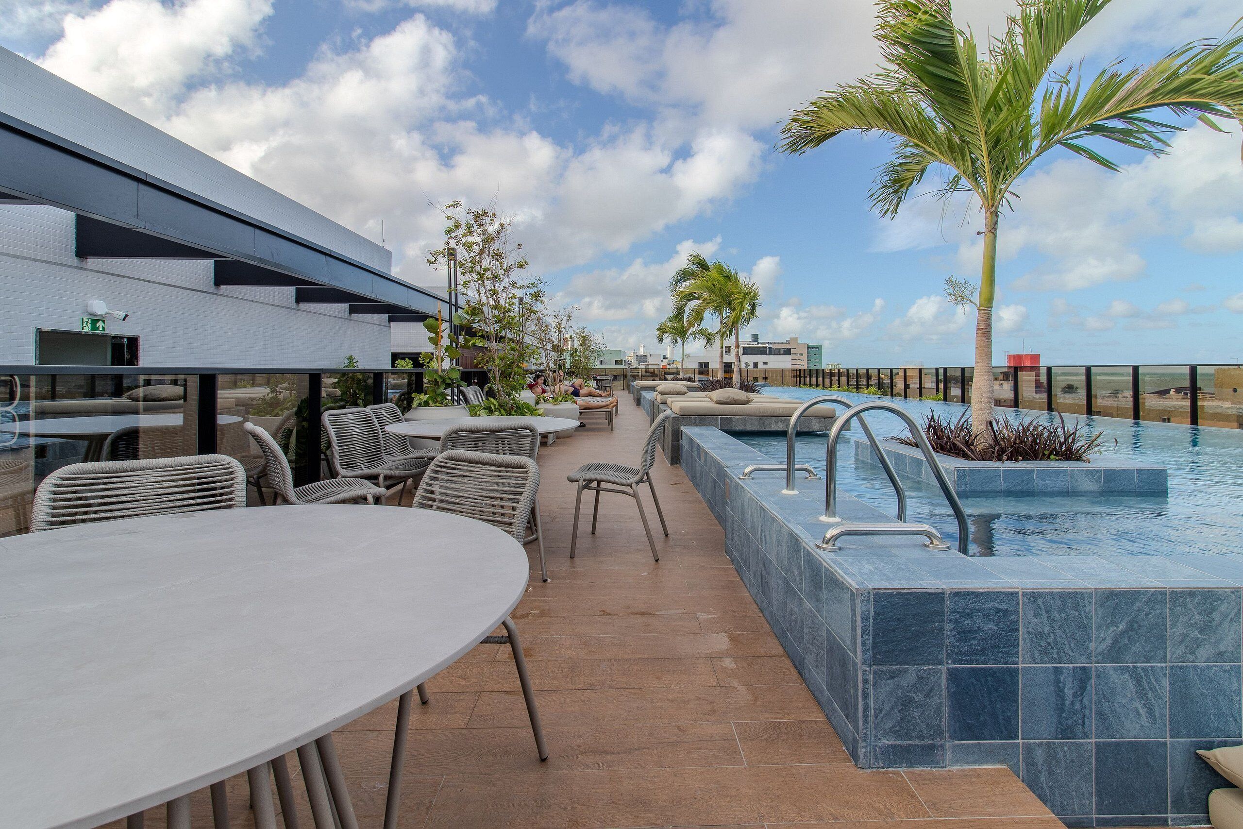 A rooftop pool area with lounge chairs, a dining table, and a view of the city skyline.