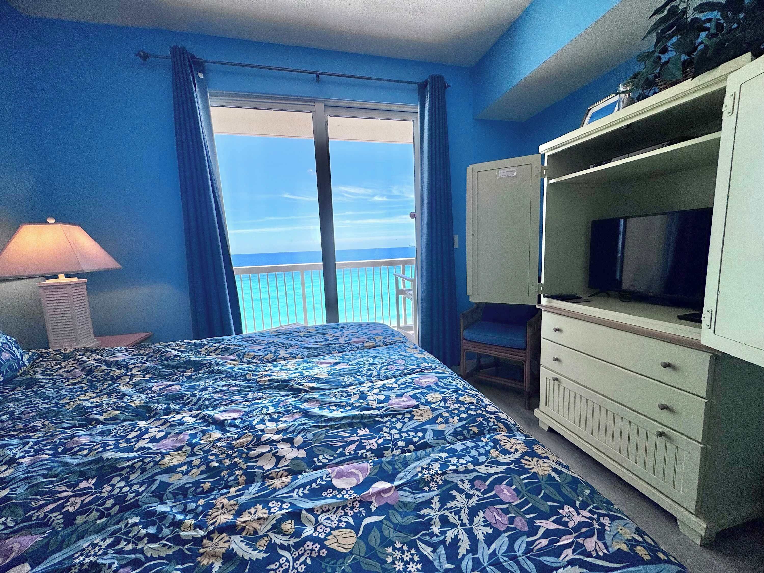 A room with a bed, a television, a dresser, and a view of the ocean.