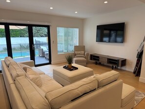 A modern living room with a beige sofa, a coffee table, a TV, and a sliding glass door leading to a pool area. - Modern Family-Friendly Retreat w/ Backyard Oasis + BBQ  Near Studios & Beaches (Los Angeles)