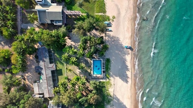 An aerial view of a beachfront property with a swimming pool, beach access, and tropical vegetation.