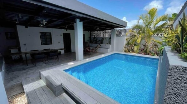 Una piscina con un porche techado, mobiliario de exterior y plantas tropicales.