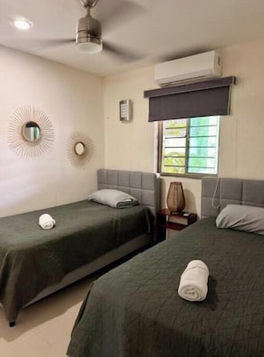 Habitación de hotel con dos camas, ventilador de techo, equipo de aire acondicionado y una ventana con persianas.