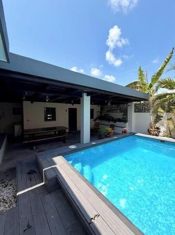 A swimming pool with a wooden deck, a covered seating area, and a tropical garden.