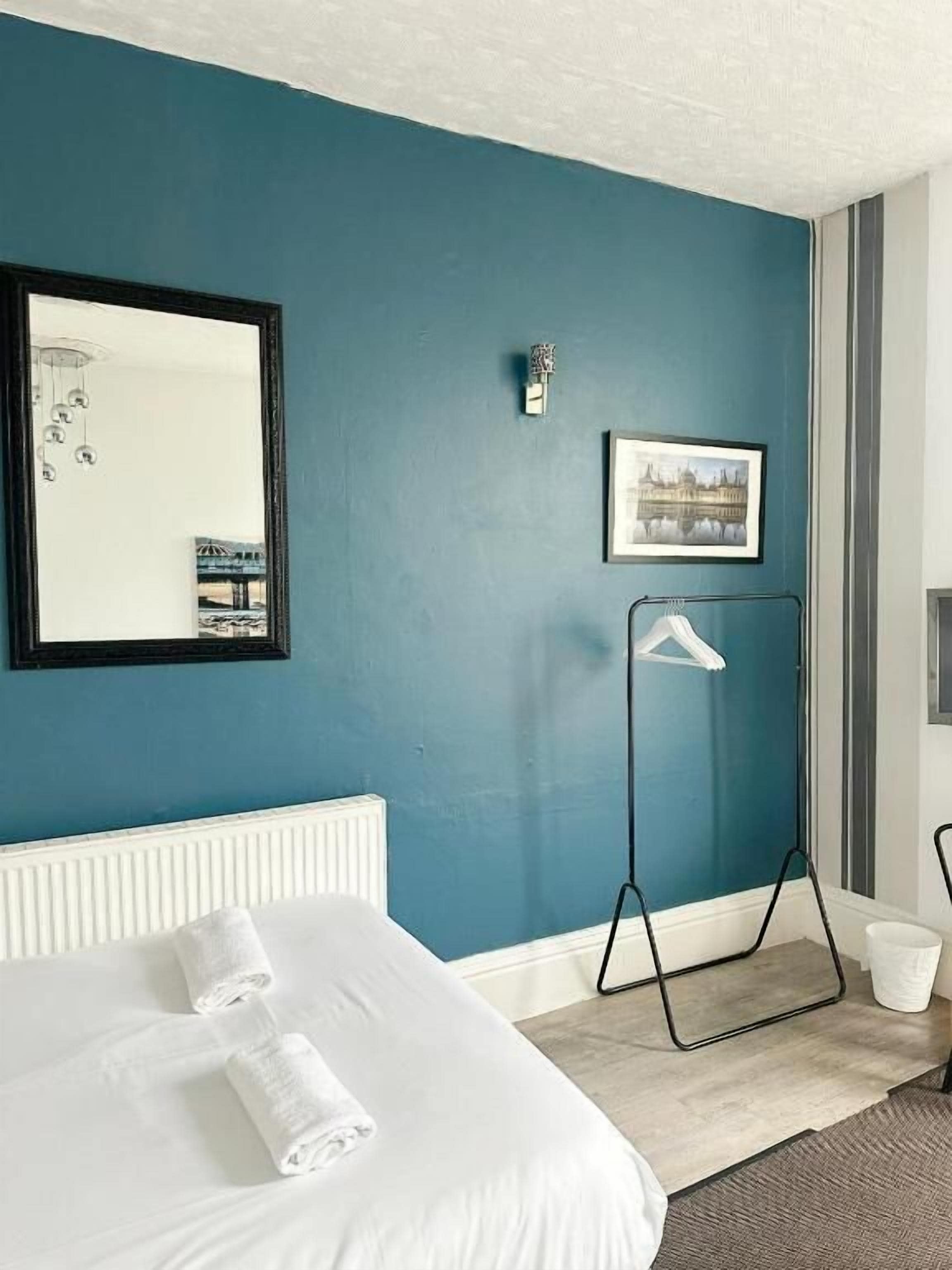 A hotel room with a bed, a radiator, a mirror, a picture frame, a hanging light fixture, and a radiator cover.
