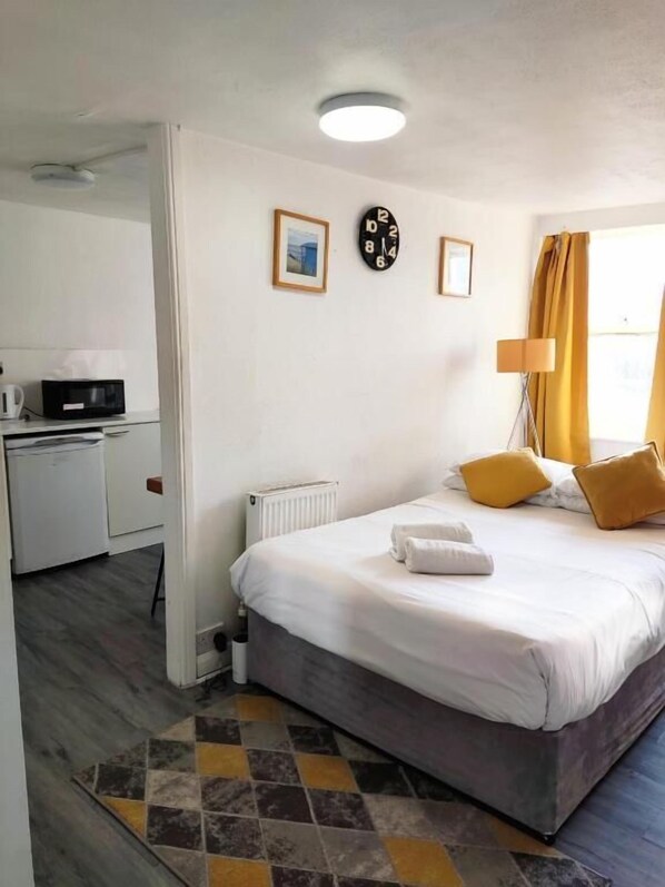Une chambre d’hôtel avec un lit, un bureau, un radiateur, une horloge et une fenêtre avec des rideaux.