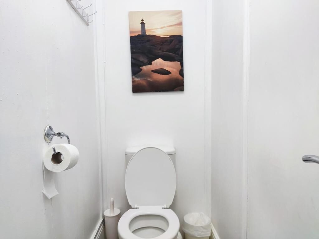 A bathroom with a toilet, a roll of toilet paper, a trash bin, and a wall-mounted picture of a lighthouse.