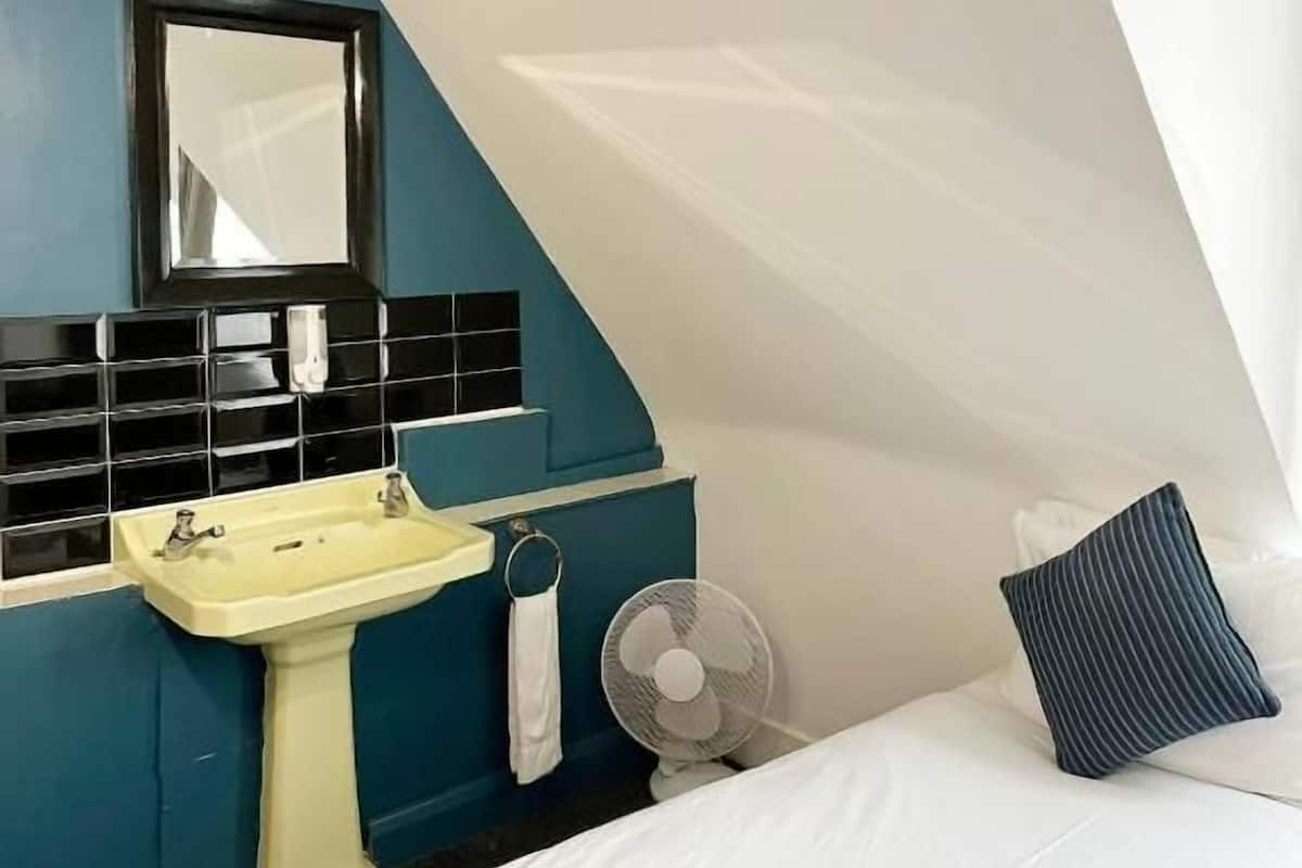 A bedroom with a bed, a bedside table, a mirror, a sink, and a fan.