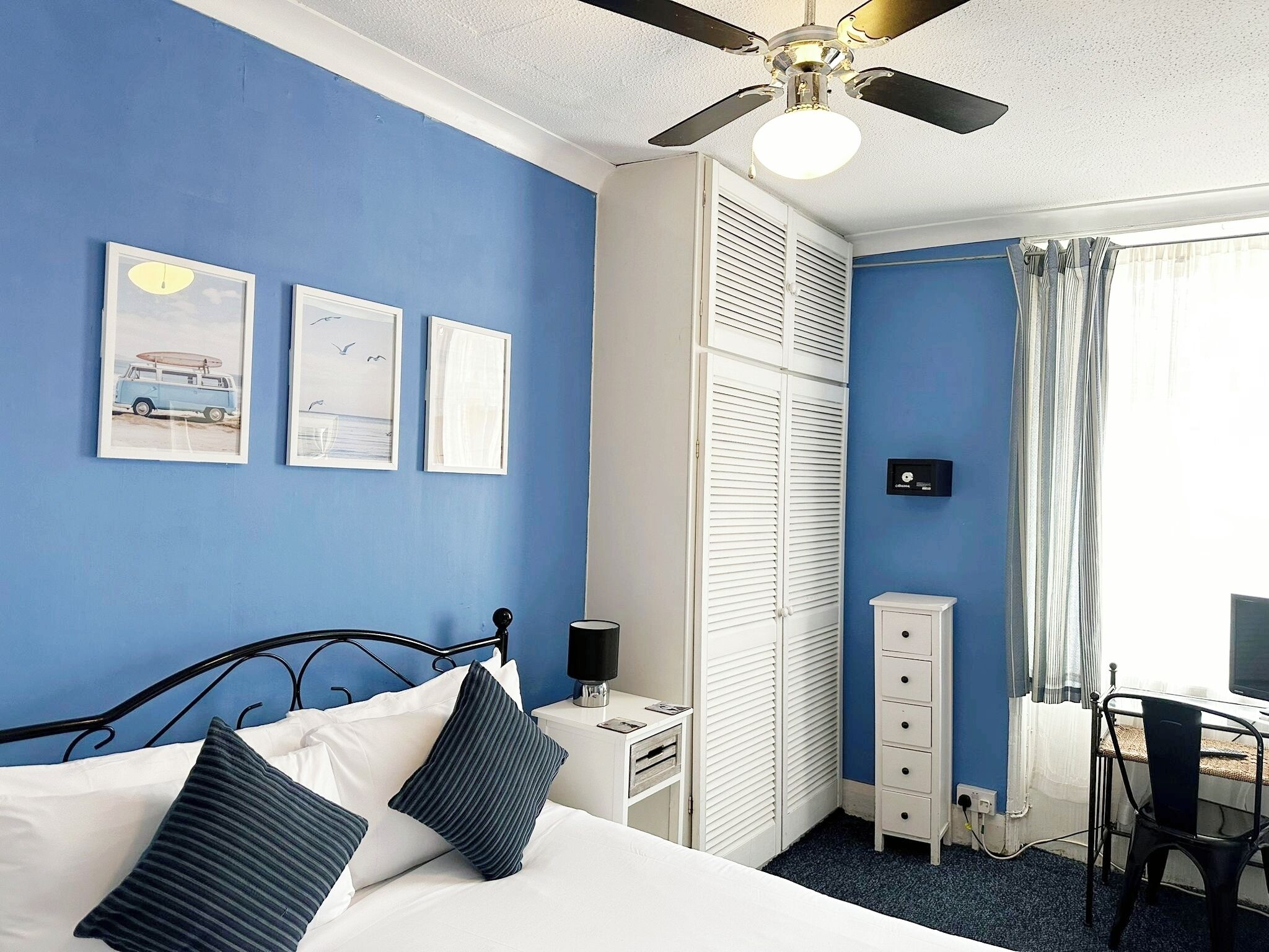 Un dormitorio con cama, escritorio, silla, ventilador de techo y ventana con cortinas.
