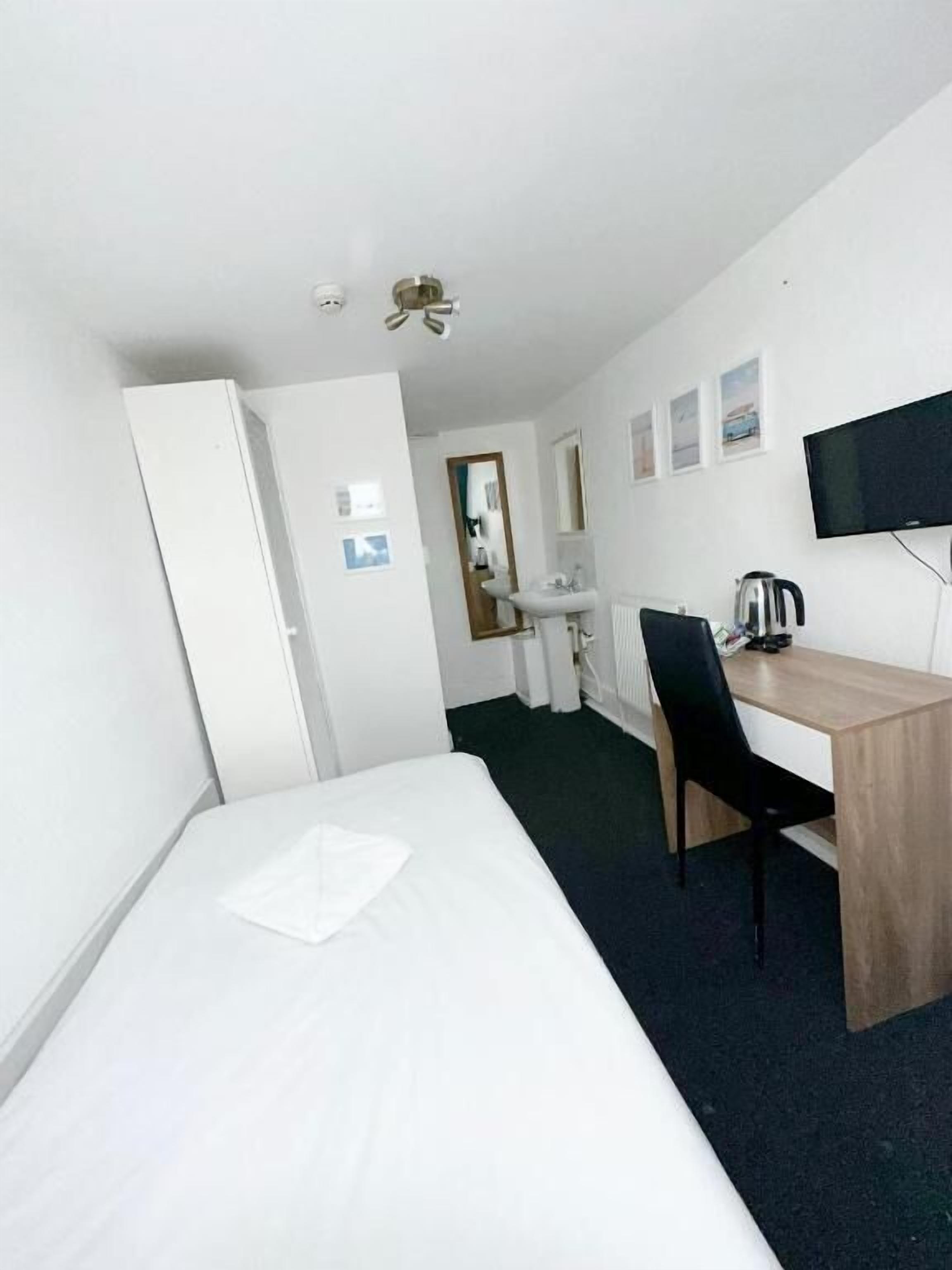 Ein Hotelzimmer mit einem Einzelbett, einem Schreibtisch mit Stuhl, einem Fernseher und einer kleinen Küchenzeile.