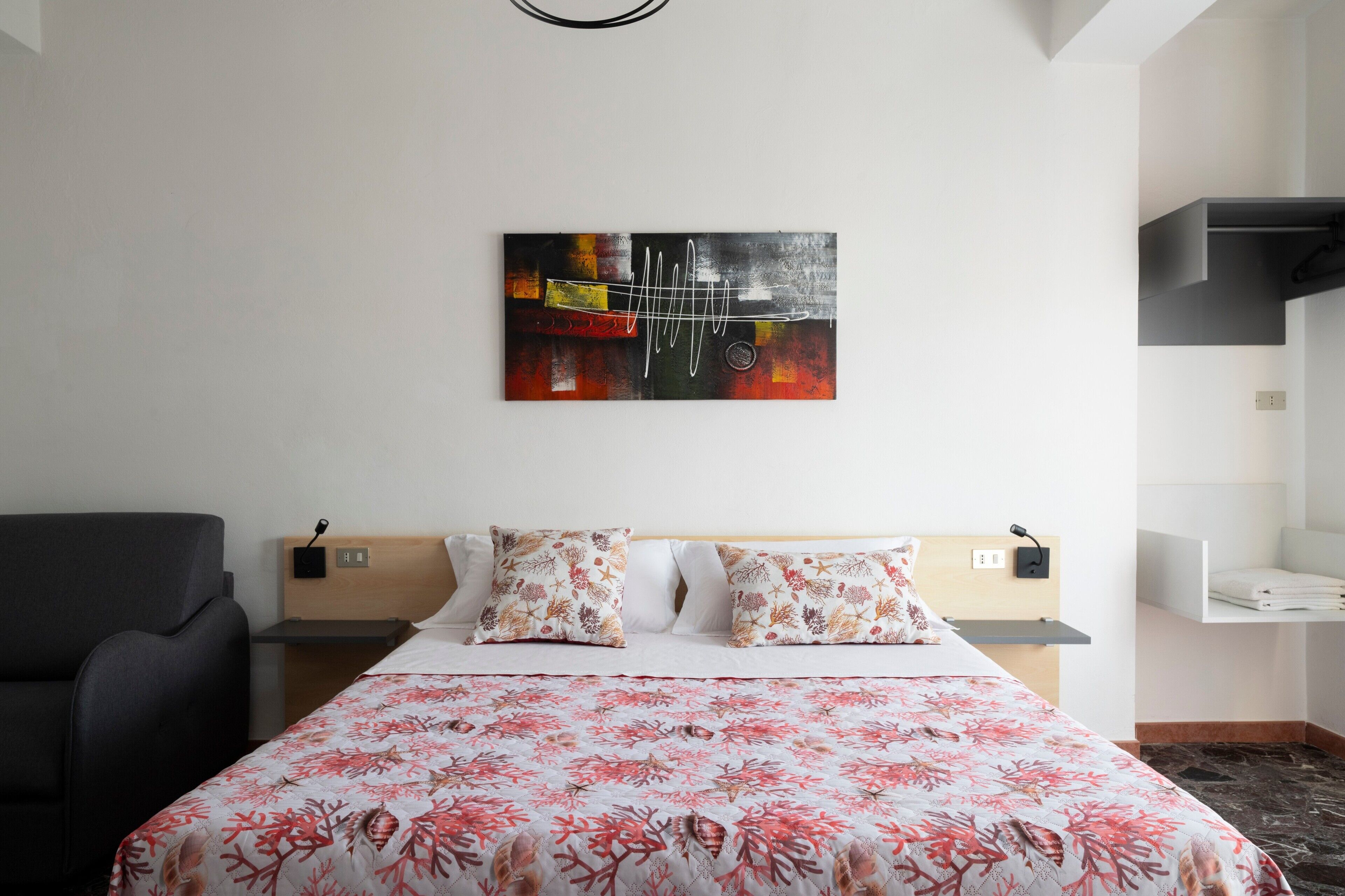 A hotel room with a bed, a chair, a nightstand, a wall-mounted artwork, and a shelf.