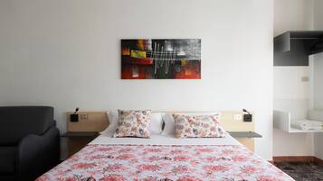A hotel room with a bed, a chair, a nightstand, a wall-mounted artwork, and a shelf.