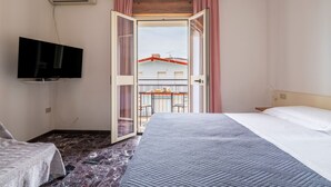Hotellihuone, jossa on sänky, seinälle asennettu televisio sekä parveke, jossa on pöytä ja tuolit.