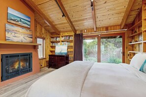 A bedroom with a fireplace, bookshelves, a bed, and a view through a glass door. - Mendocino Oceanfront Home - Hot Tub & BBQ (Mendocino)