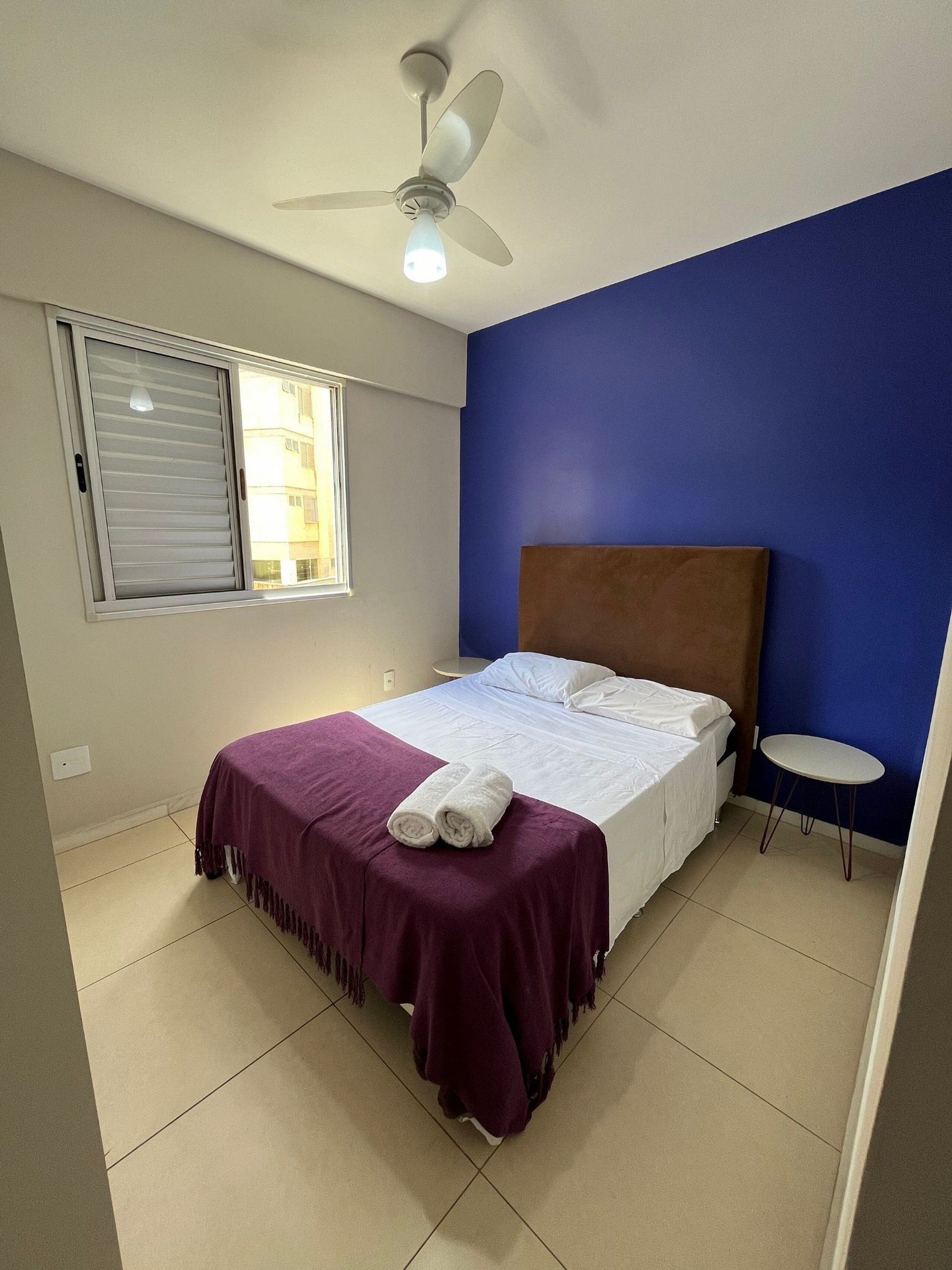 Ein Schlafzimmer mit einem Bett, einem Deckenventilator, einem Fenster mit Jalousien und einem kleinen runden Tisch.