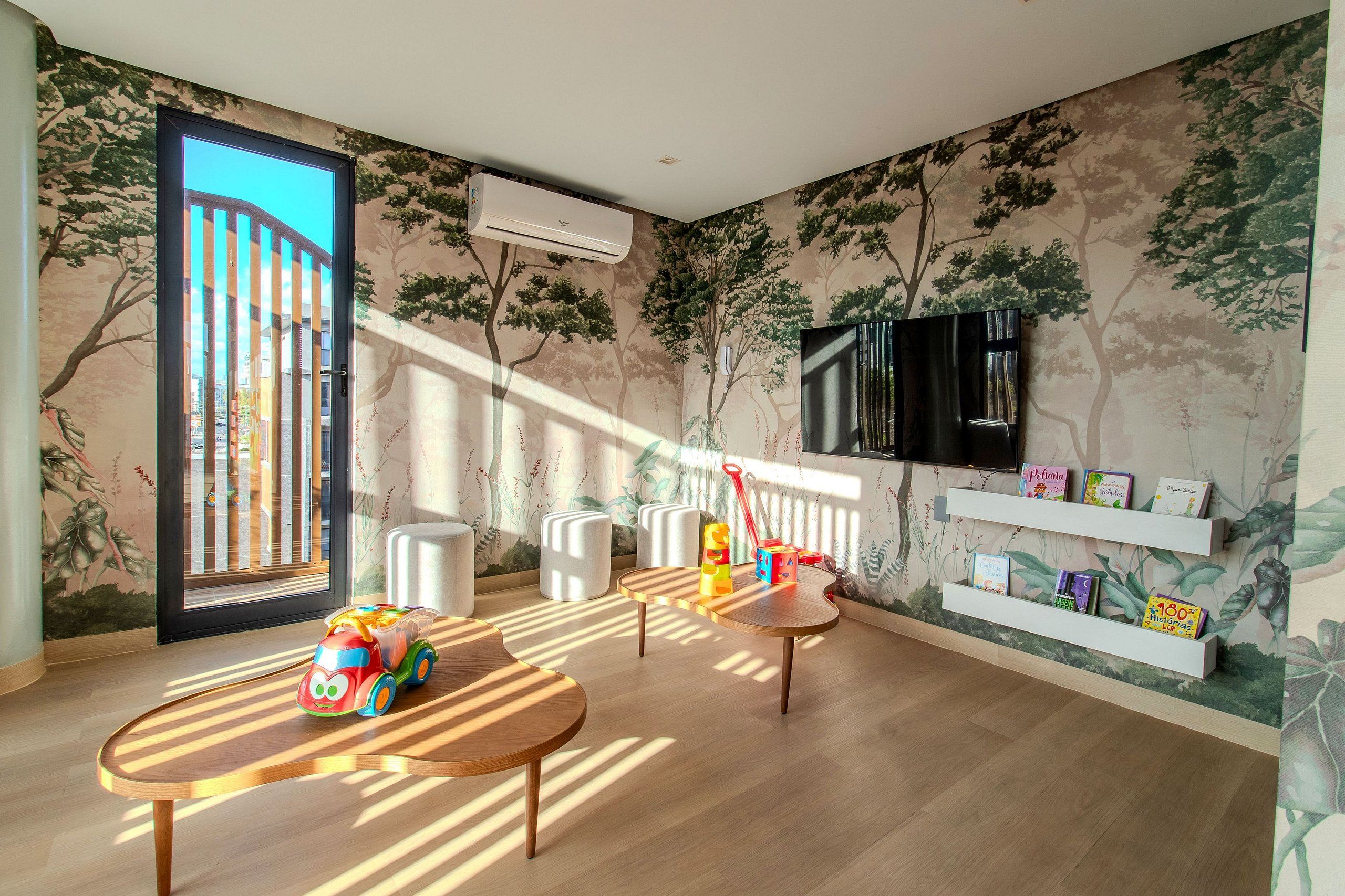 A room with a large window, a wall-mounted TV, and a shelf with children's books.