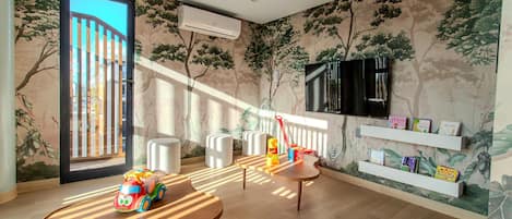 A room with a large window, a wall-mounted TV, and a shelf with children's books.