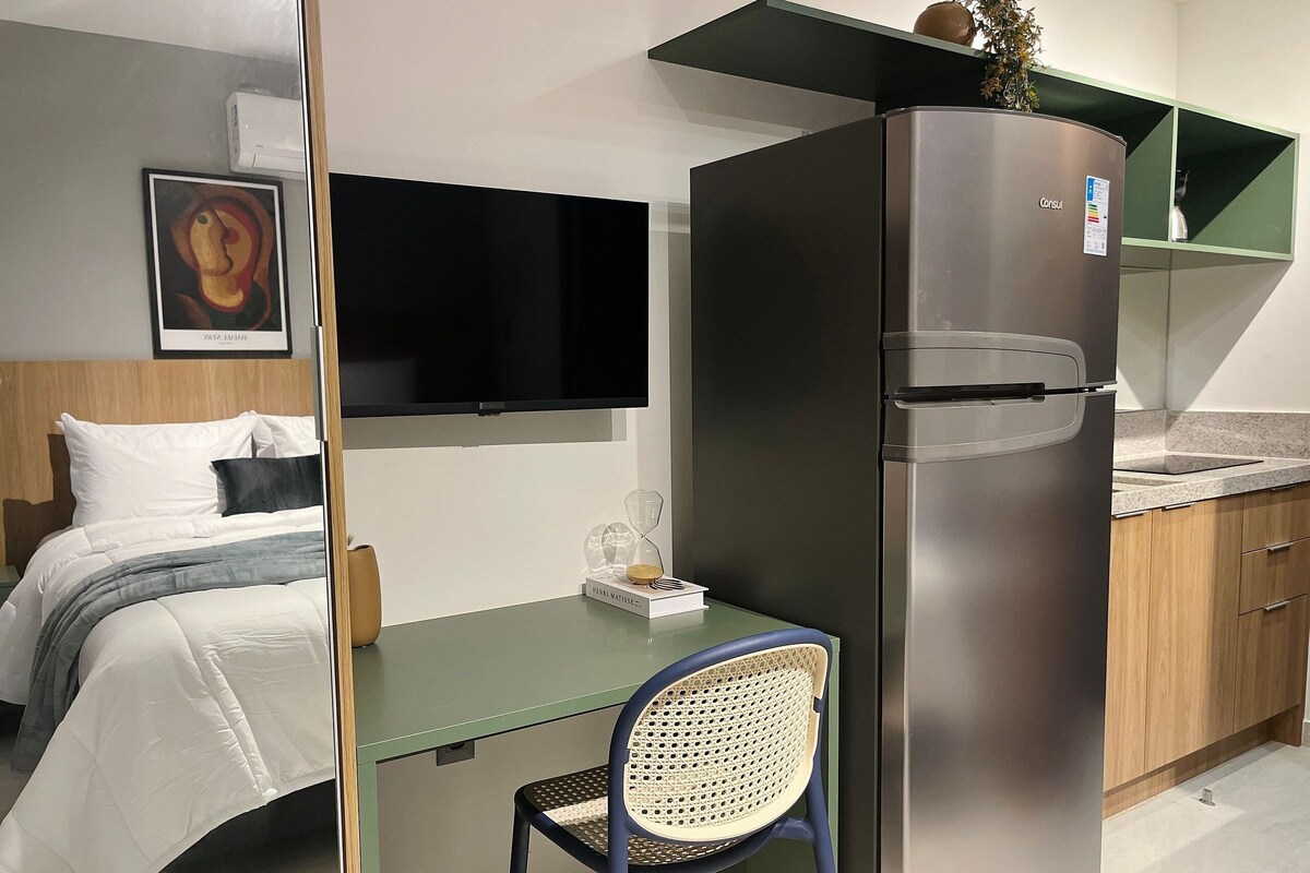 Habitación de hotel pequeña y funcional, con cama, mesita, silla, refrigerador, televisor y kitchenette.