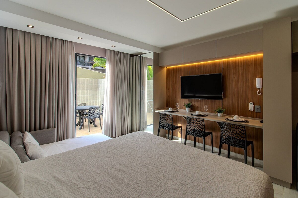 Habitación de hotel moderna con una cama grande, un escritorio con sillas, un televisor de pantalla plana y un balcón con cortinas.
