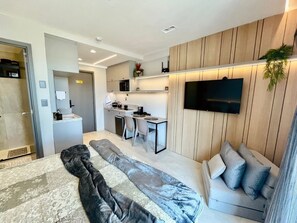 Una habitación de hotel moderna con cama, sofá, escritorio con silla, televisor montado en la pared y pequeña zona de cocina.