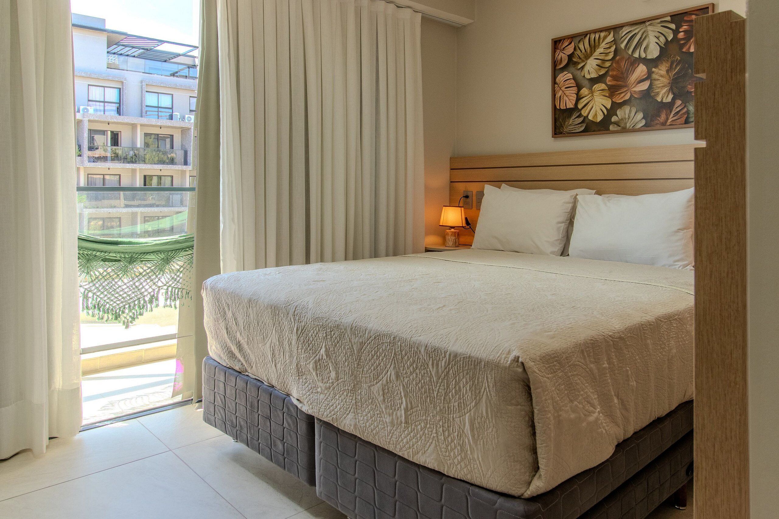 Habitación de hotel con cama, cortinas, mesita de noche con lámpara, pared con diseño de hojas y vista a un edificio.