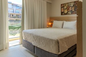 A hotel room with a bed, curtains, a nightstand with a lamp, a wall with a leaf pattern, and a view of a building outside. - Garden Haus (João Pessoa)