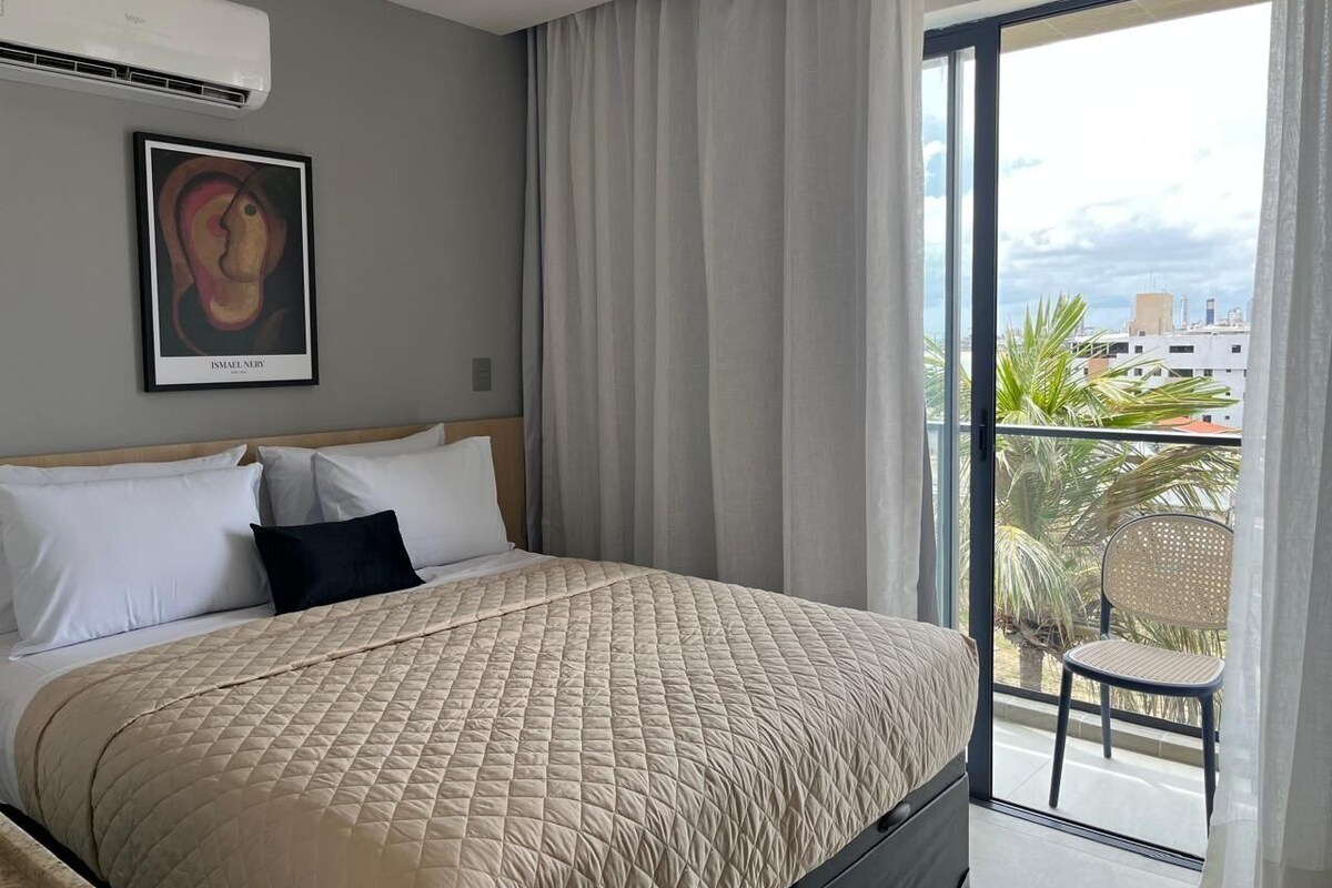 Habitación de hotel con cama, una obra de arte enmarcada, un balcón con una silla y vistas a edificios y palmeras.