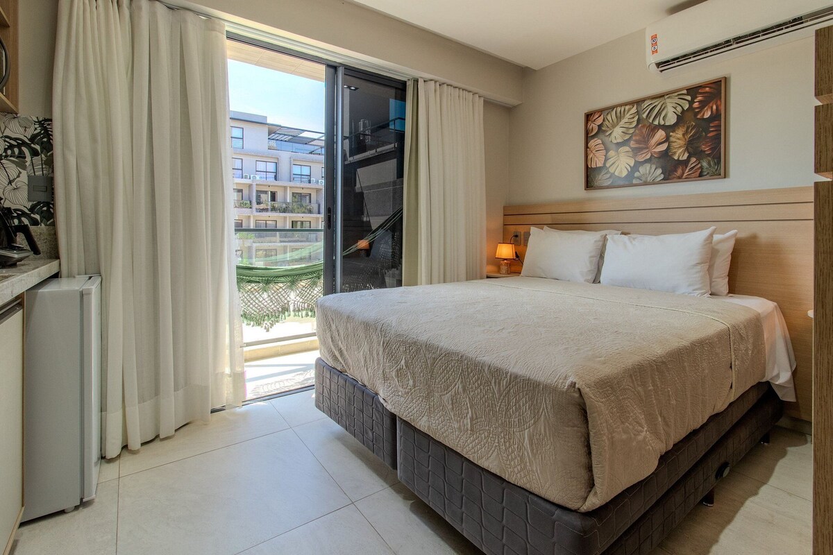 Habitación de hotel con cama, escritorio, silla, ventana con cortinas, un cuadro en la pared y balcón con vistas a edificios.