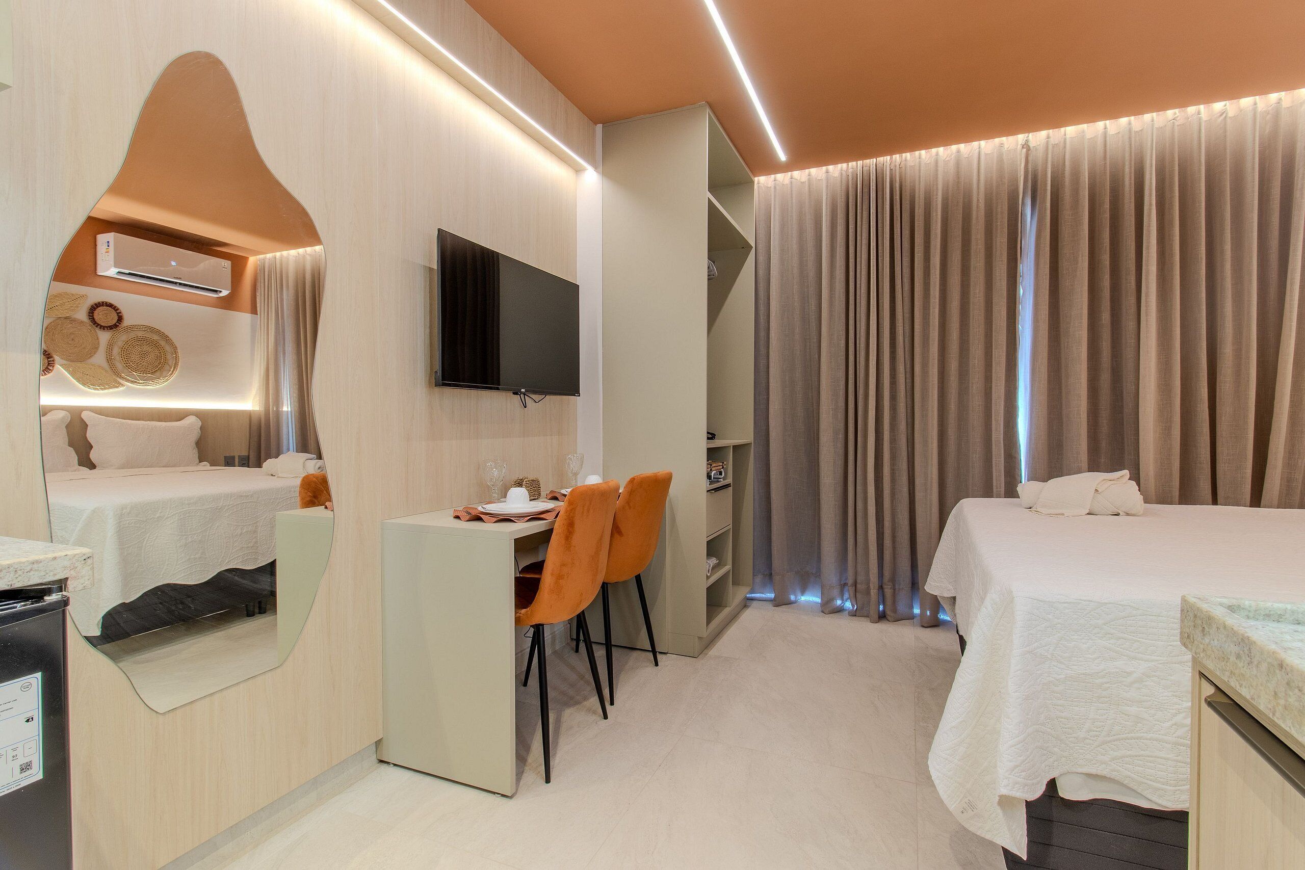 Quarto de hotel com uma cama, uma escrivaninha com cadeiras, uma televisão e um espelho.