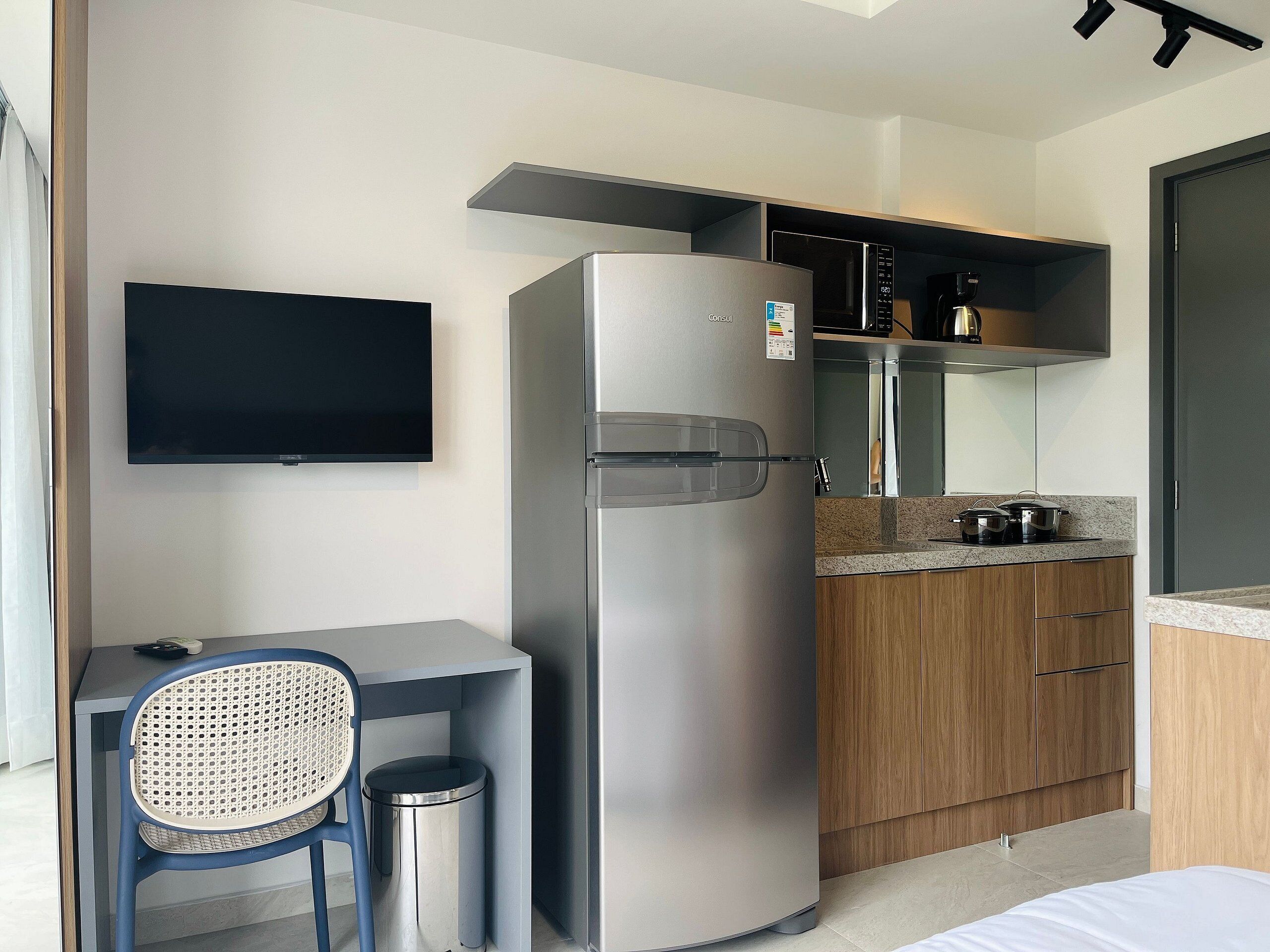 Una kitchenette moderna con refrigerador, microondas y estufa integrados, una pequeña zona de comedor con una silla y mesa, y un televisor montado en la pared.