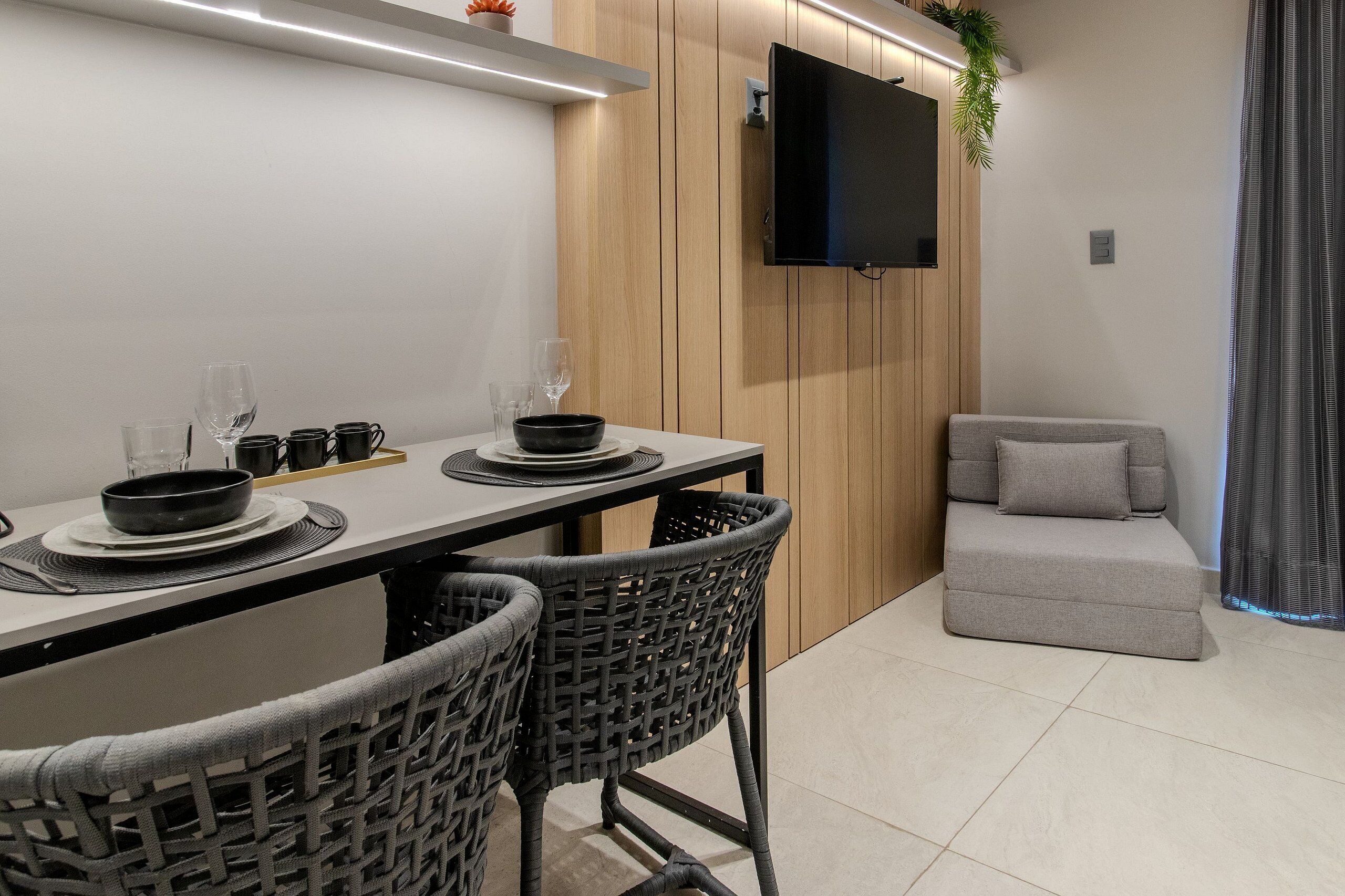 Uma zona de refeições moderna com uma mesa posta para quatro pessoas, rodeada por cadeiras de vime, um sofá cinzento e uma televisão de ecrã plano montada numa parede de madeira.