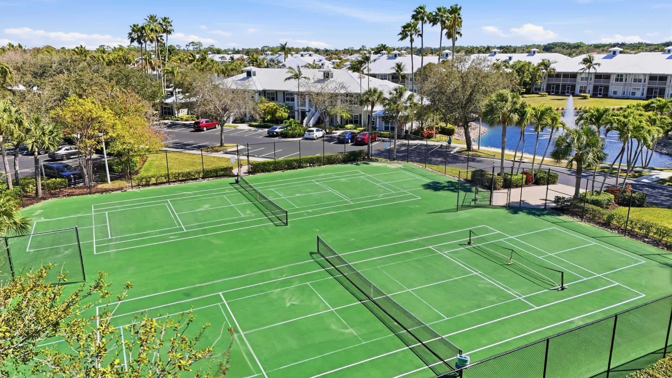 A tennis court with a net, surrounded by a fence, with a parking lot and buildings in the background.