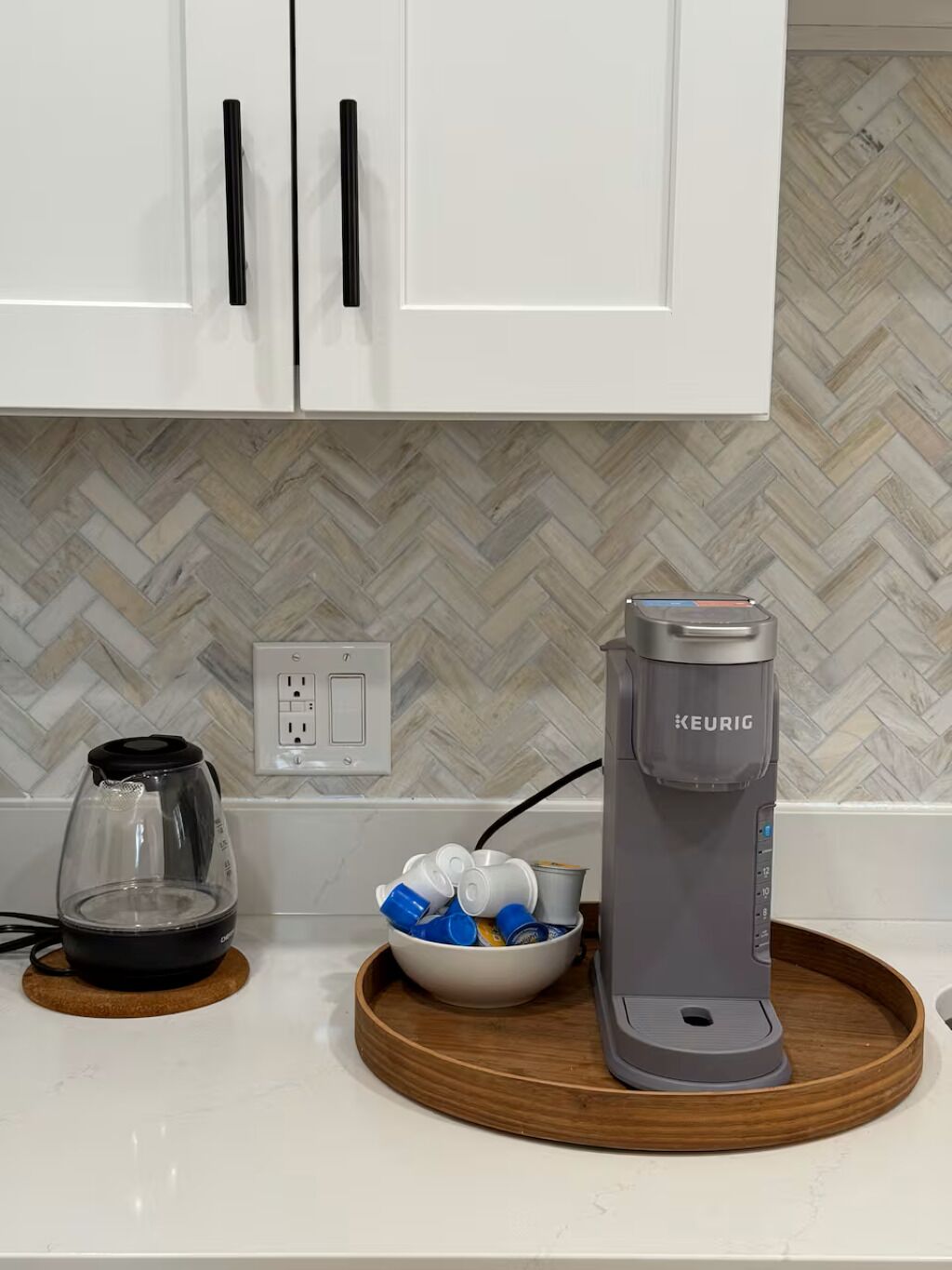 Une cuisine avec des meubles blancs, un pare-feu décoré d’un motif en chevrons, une machine à café, une carafe en verre et un bol rempli de tasses, le tout posé sur un plateau en bois.