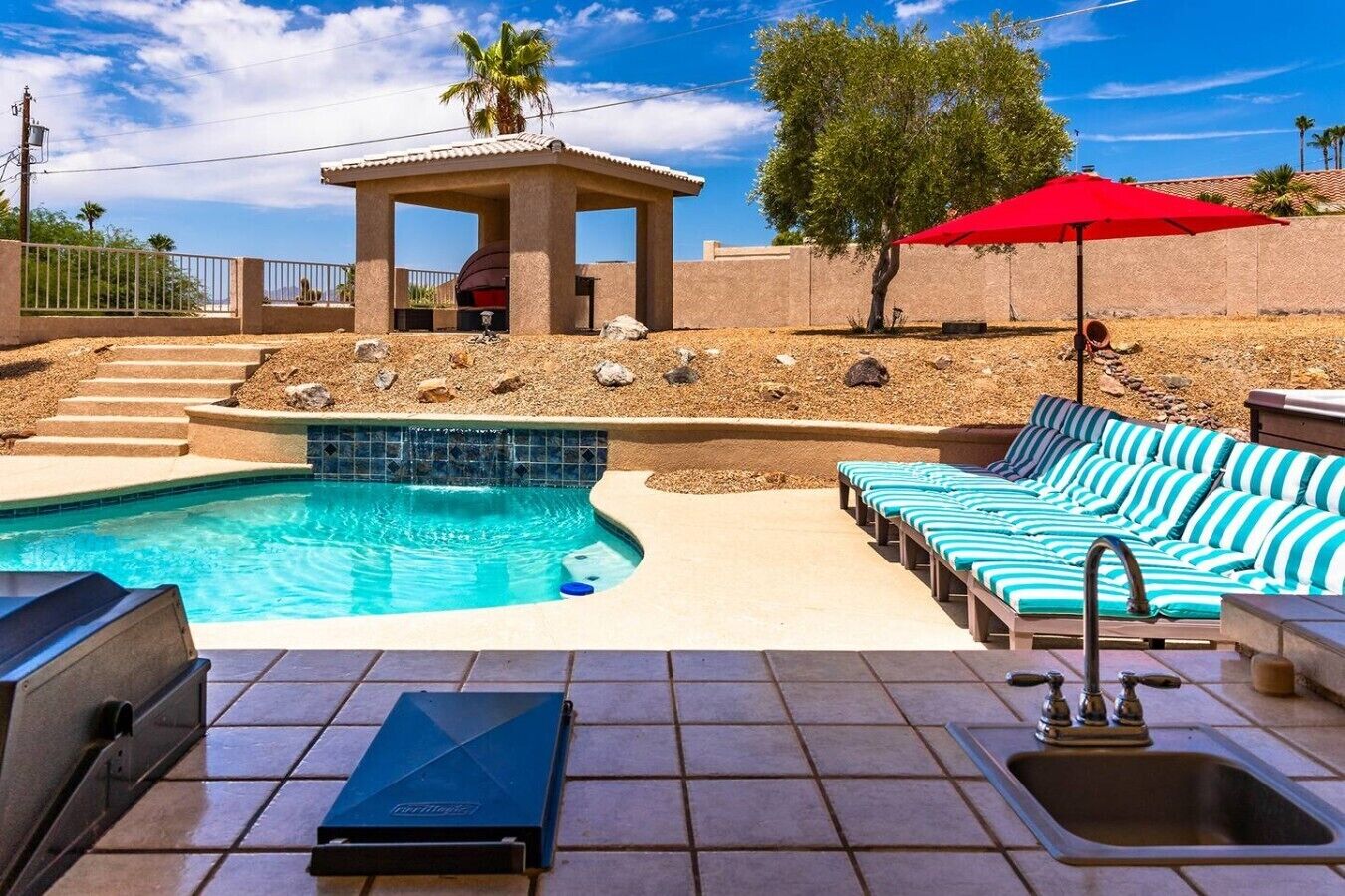 Une piscine avec des chaises longues, un jacuzzi et une pergola surmontée d’un parasol rouge.