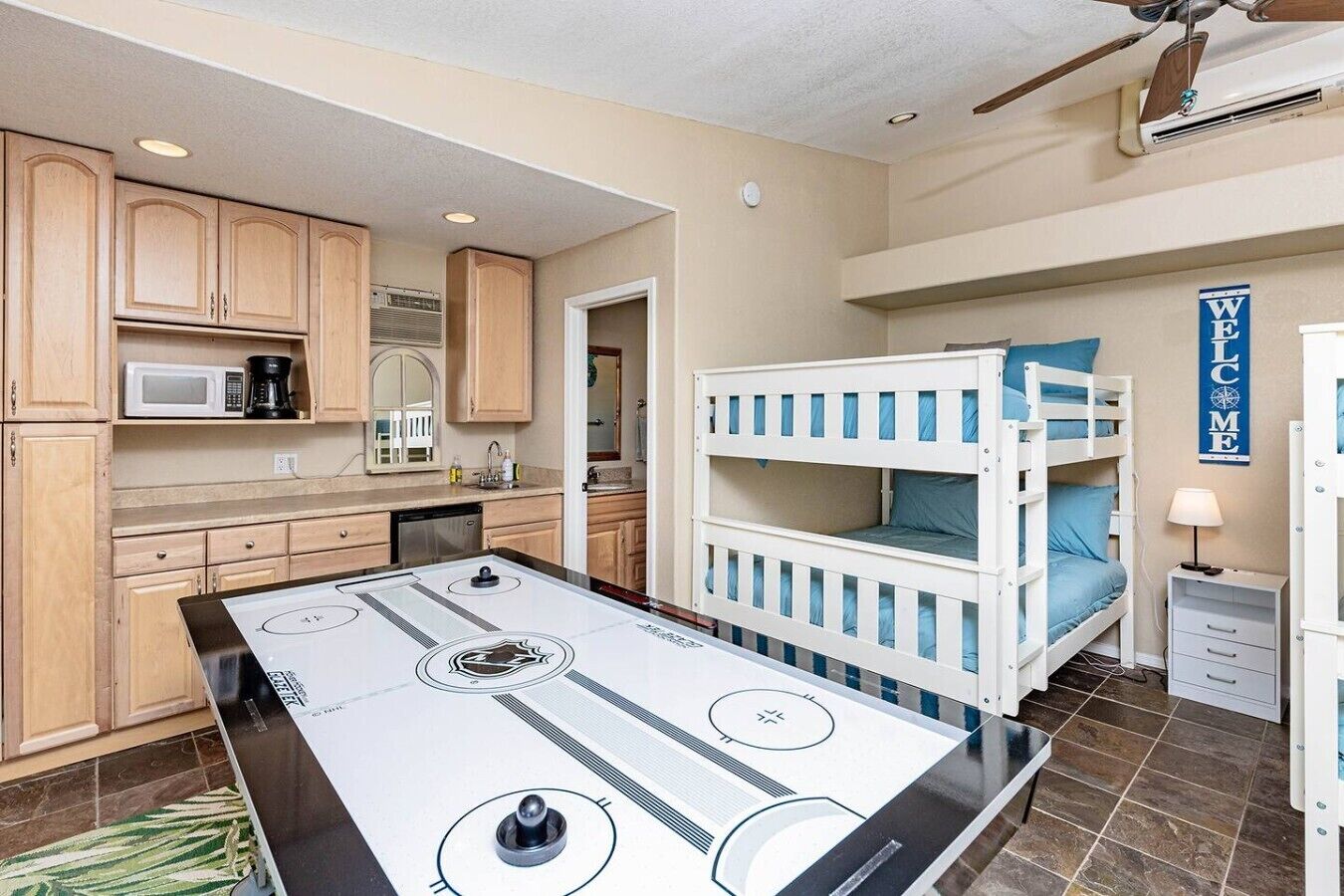 A kitchen with wooden cabinets, a microwave, and a coffee maker. There is a bunk bed with blue bedding and a welcome sign.