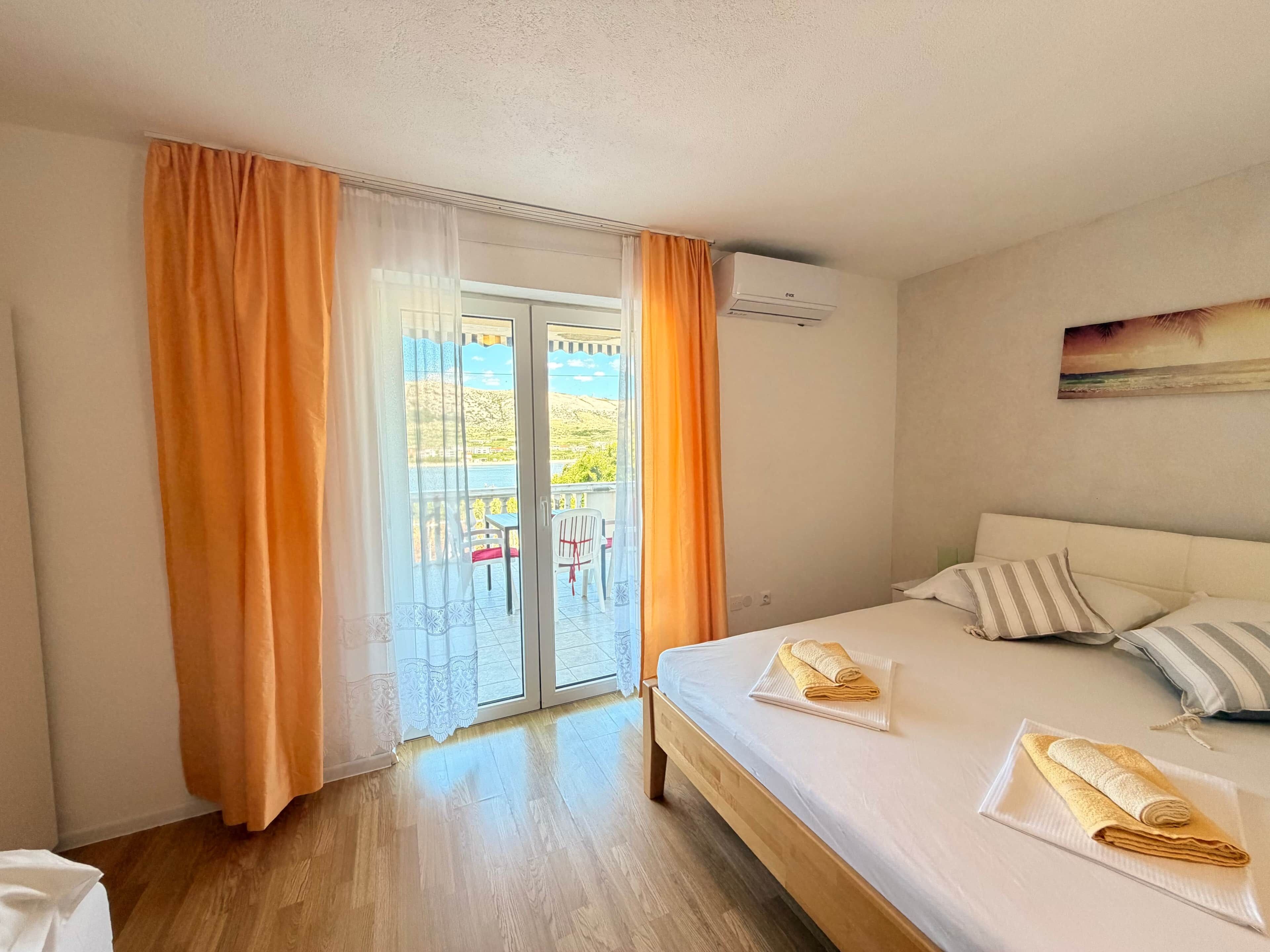A hotel room with a bed, a balcony with orange curtains, and a view of a landscape.