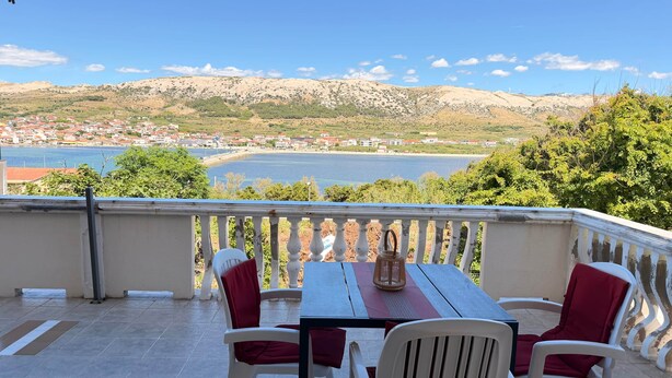 En balkon med et bord og stole, der udover et kystlandskab med bygninger og bakker.